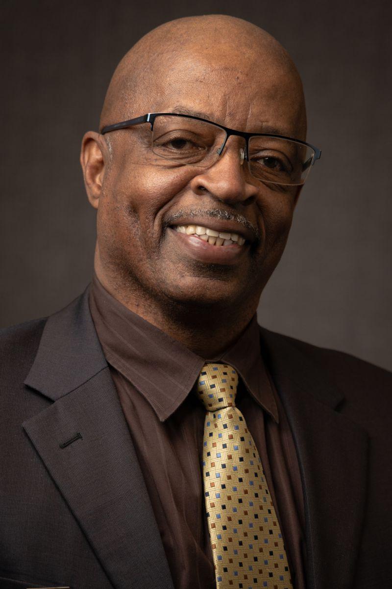 Harold Wright, an African American male wearing a grey suit jacket with a maroon collared shirt, a gold tie with black dots, and glasses.