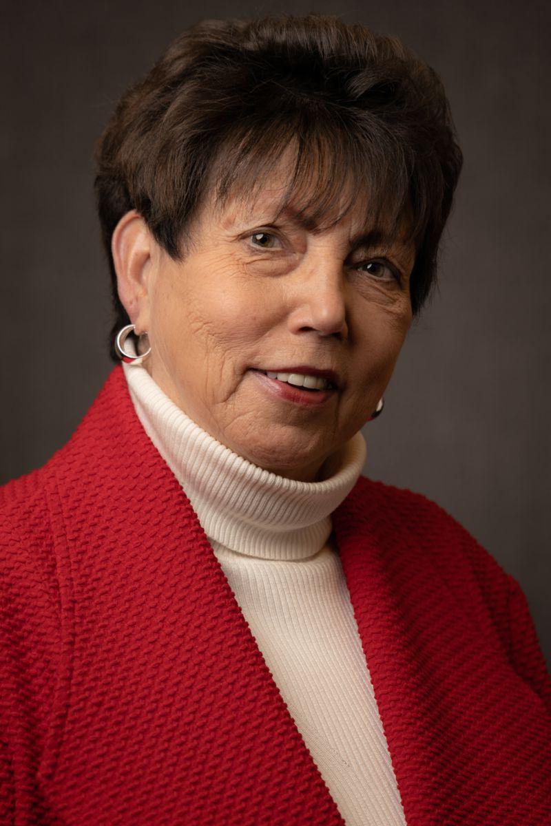 Pat Goodwin, a Caucasian female with short brown hair wearing gold earrings, a cream sweater, and a red cardigan.