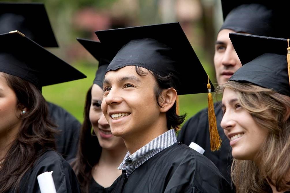 A forest green, hyperlinked, button labeled Associate Degrees in white, bolded, underlined text with an image of six graduates smile to the right of the frame in black graduate caps and gowns: A Caucasian female with sandy, brown hair stands to the right of a smiling, brown haired, Caucasian male with a blue collared shirt showing above his gown. To his left a brown haired, Caucasian female stands smiling with another brown haired, Caucasian female standing partially out of the frame to her left. A Caucasian male stands partially blocked by the first female and a sixth graduate stands hidden behind the last female.